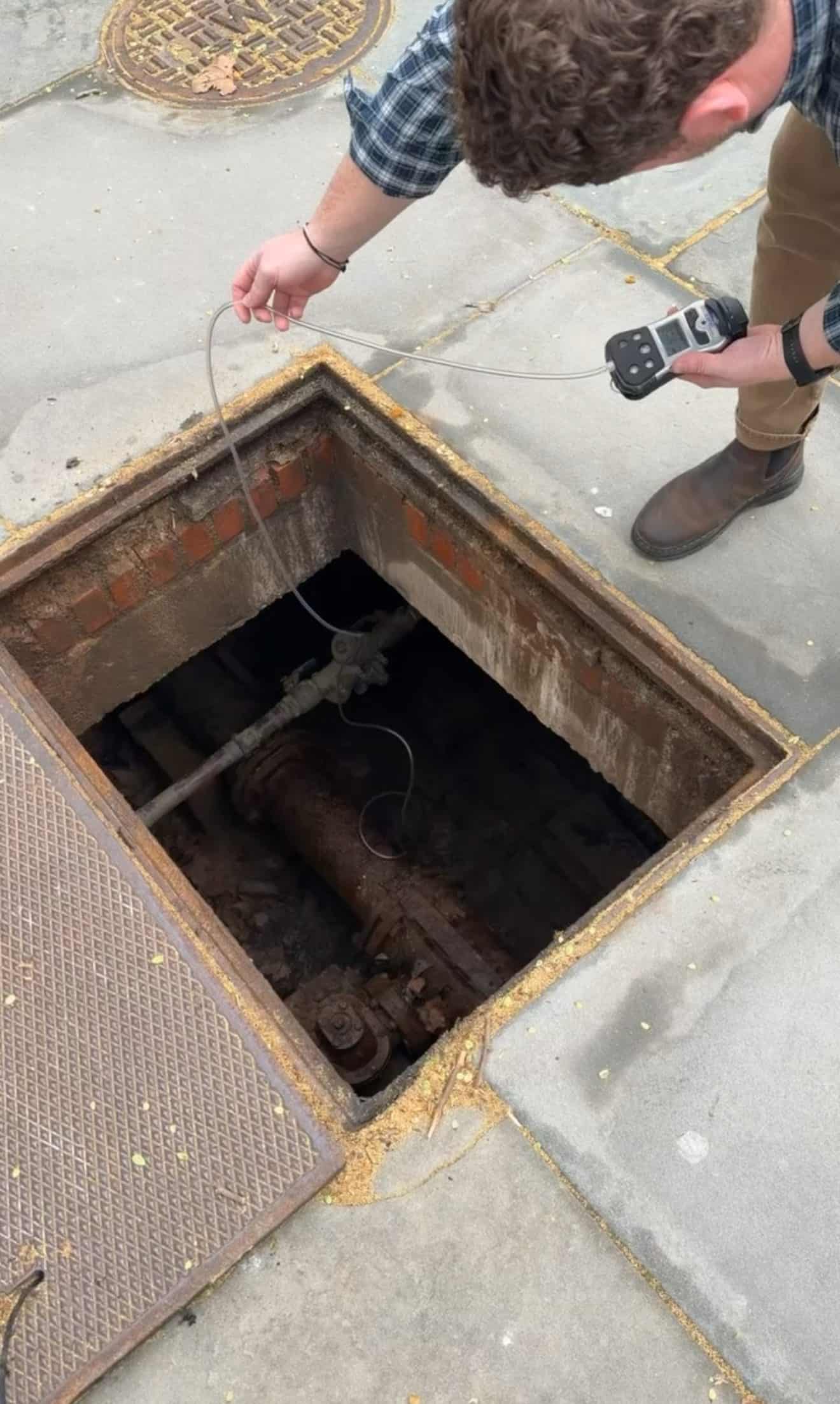 Technician lowering a sampling line from a four-gas meter into an open below-grade vault to test the atmosphere before entry