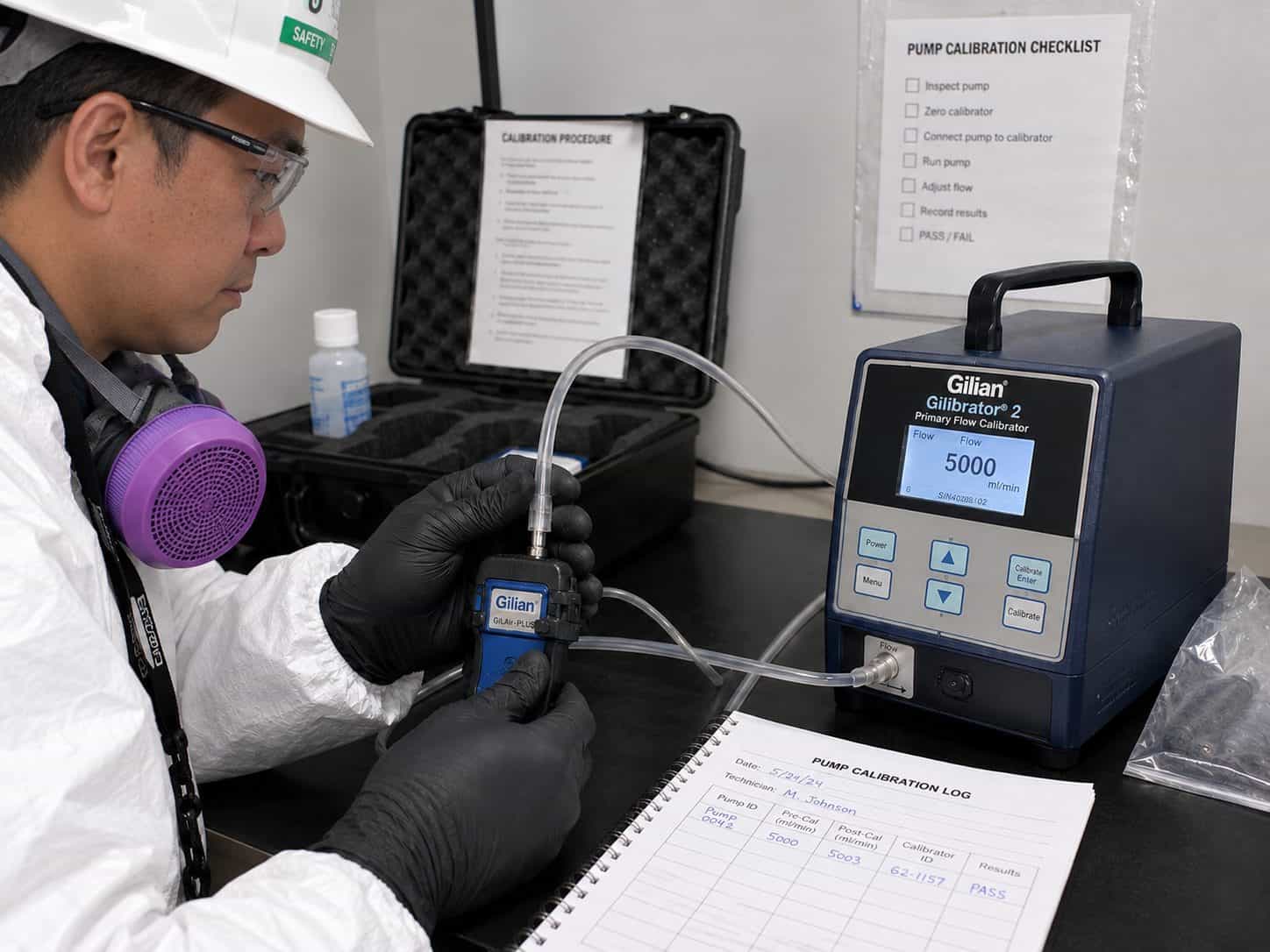 Technician in protective coveralls calibrating a personal air sampling pump against a primary flow calibrator, with the calibration log alongside